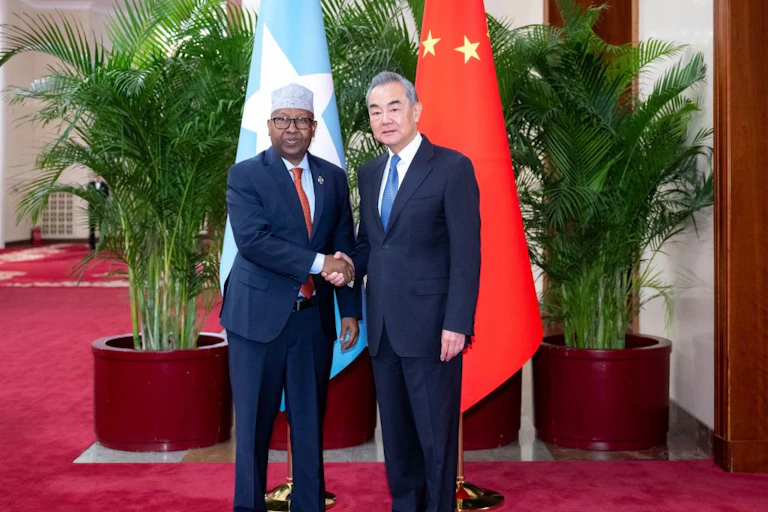 Two men looking at the camera while shaking hands and standing in front of blue and red flags
