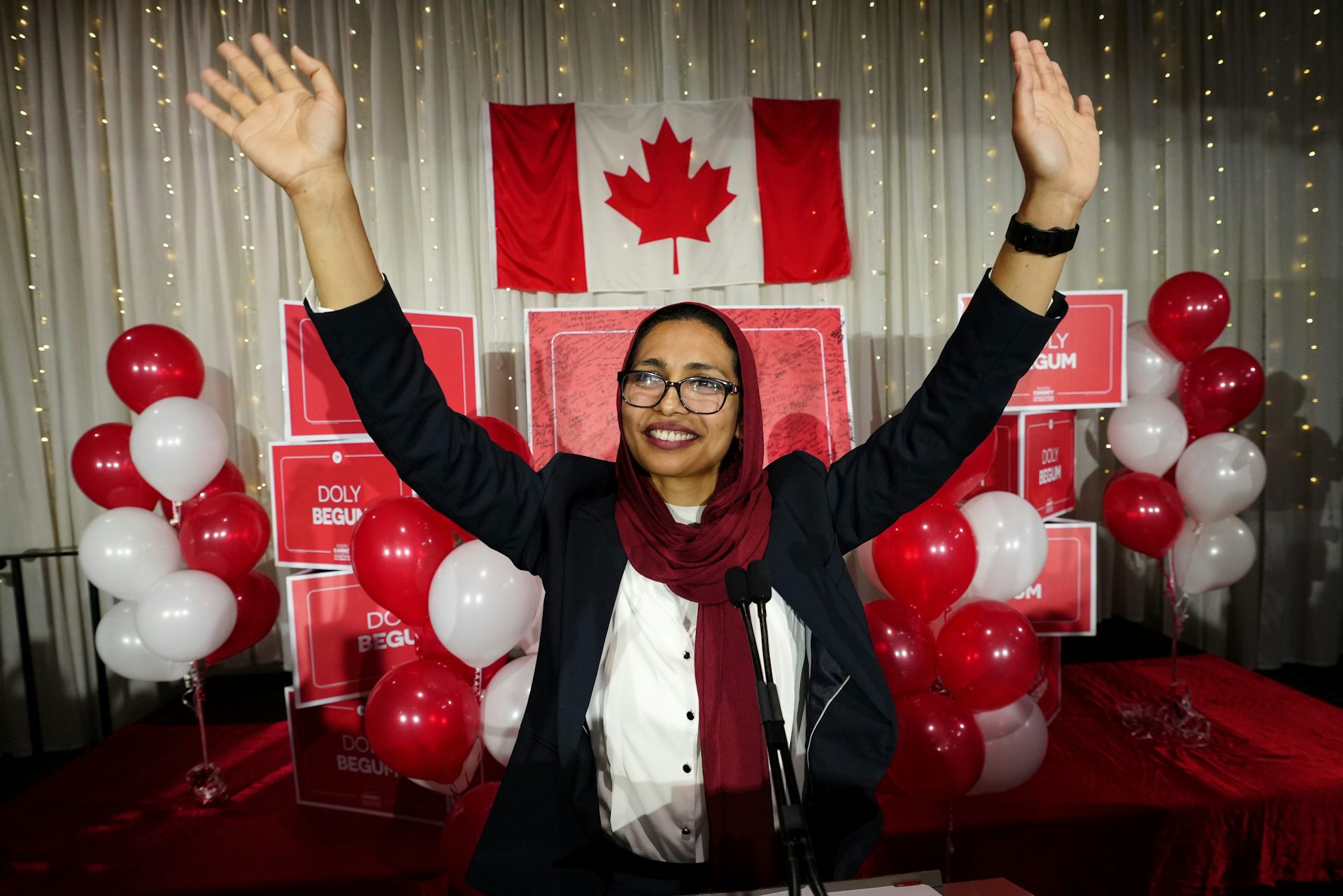A smiling woman in a headscarf raises her hands in front of a canadian flag