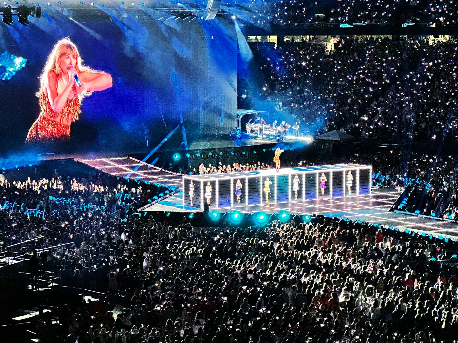 A giant image of Taylor Swift on a screen looms over a concert crowd while she performs on stage during her Eras tour in Foxborough, Mass.
