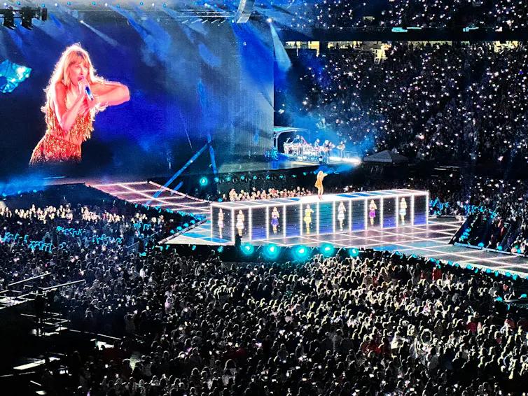 A giant image of Taylor Swift on a screen looms over a concert crowd while she performs on stage during her Eras tour in Foxborough, Mass.