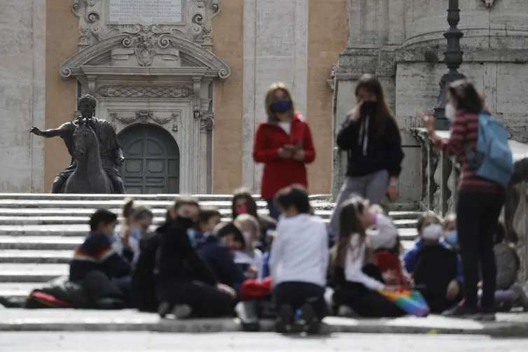 Jóvenes con un adulto visto al aire libre, borrosos sobre una estatua al fondo.
