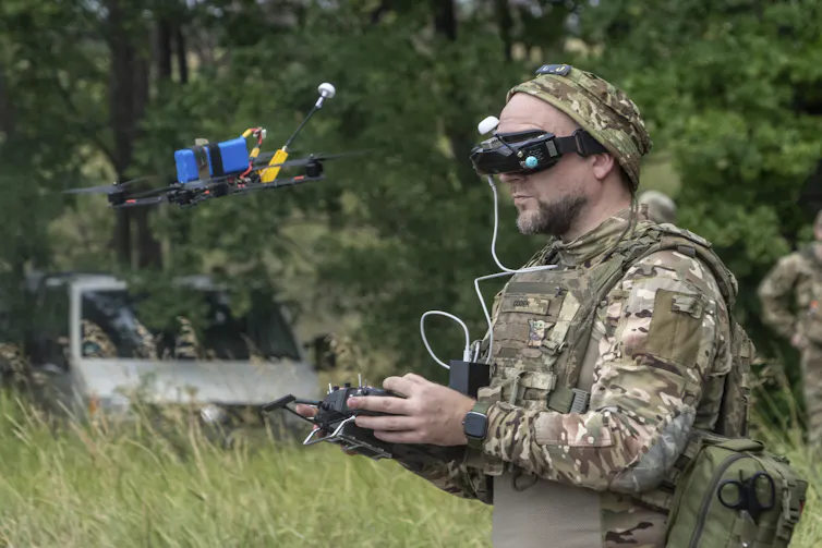 a man in military clothing and wearing goggles holds a device in his hands as a quadcopter hovers in front of him