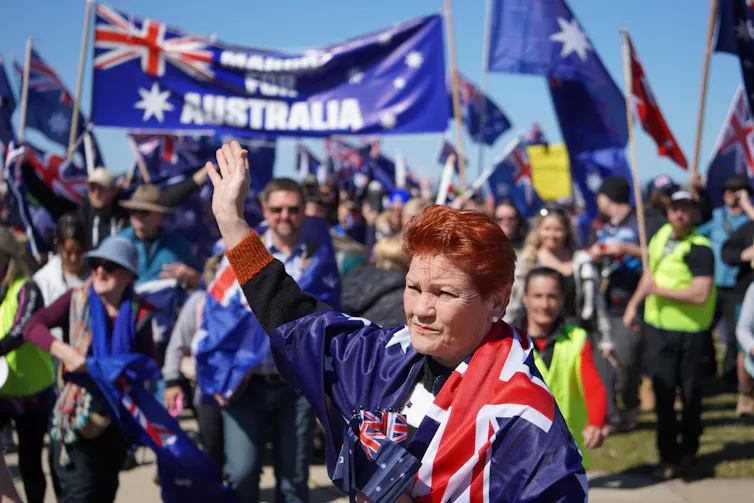 Pauline Hanson wrapped in an Australian flag, in front of a rally, with Australian flags