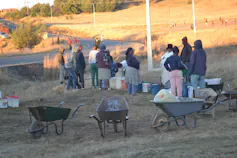 A line of people with water containers and a wheelbarrow queuing for water on the side of a road