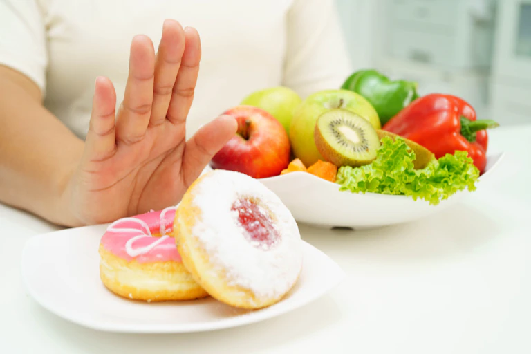 Hand pushes away a plate of two donuts. A bowl of fruit and salad is in the background.