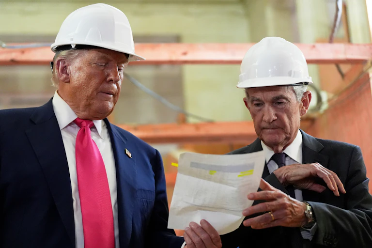 Two white men wearing hard hats examine a piece of paper in their hands at a construction site