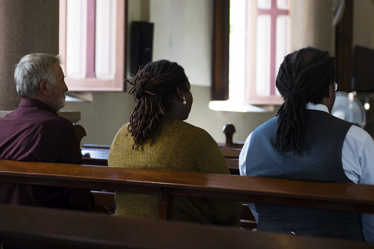 Three adults sit in a church pew, with windows with cross decorations in the background.