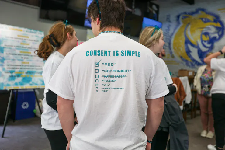 A person wears a white shirt that says 'Consent is simple' with a checkmark box below it that is checked and says 'yes,' as well as other words like 'Not Tonight' crossed out.