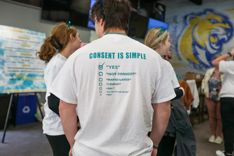 A person wears a white shirt that says 'Consent is simple' with a checkmark box below it that is checked and says 'yes,' as well as other words like 'Not Tonight' crossed out.