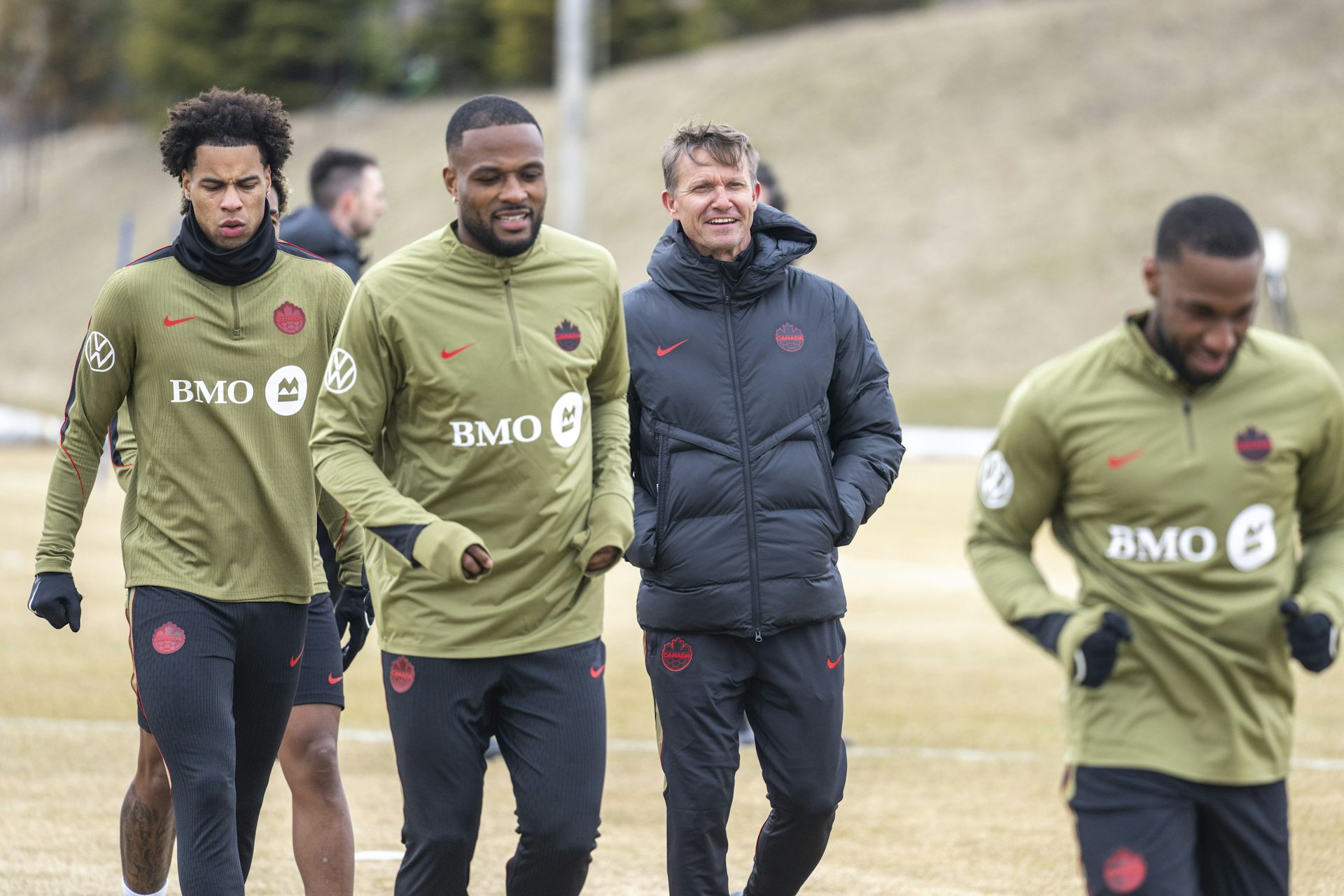 Un hombre blanco de mediana edad con una parka camina por el campo, con tres jugadores de fútbol negros con mangas largas de color verde oliva.