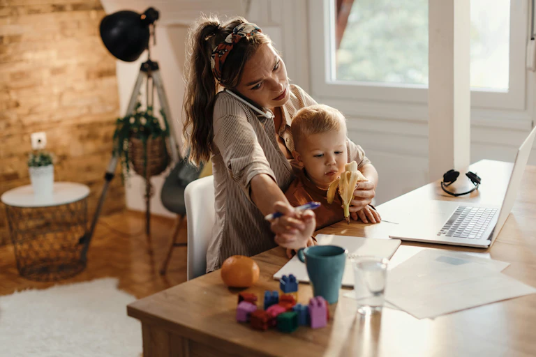 A woman is sitting at a desk facing a laptop while juggling speaking on the phone and caring for a child that is seated on her lap.