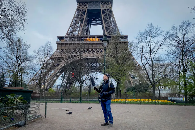 The researcher stands holding out a large black microphone, with the Eiffel Tower behind him.