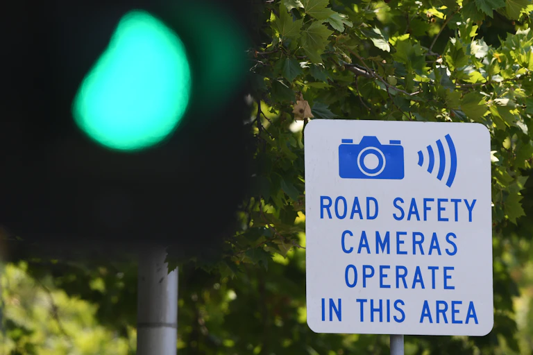 A sign for road safety cameras between a tree and a green traffic light.