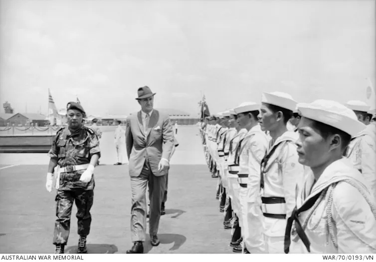 A man in a suit and hat walks next to a military commander and inspects young soldiers
