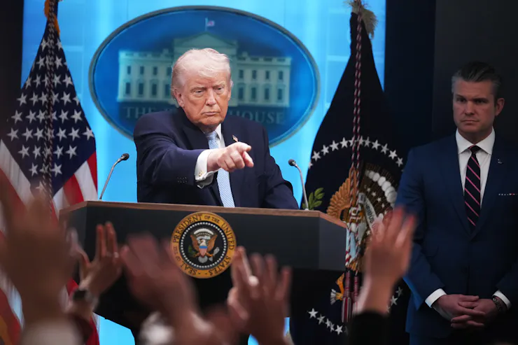 A man standing at a lectern flanked by flags, pointing into the audience of raised hands.