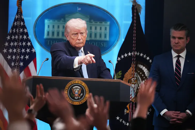 A man standing at a lectern flanked by flags, pointing into the audience of raised hands.