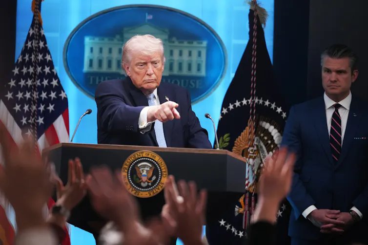 A man standing at a lectern flanked by flags, pointing into the audience of raised hands.