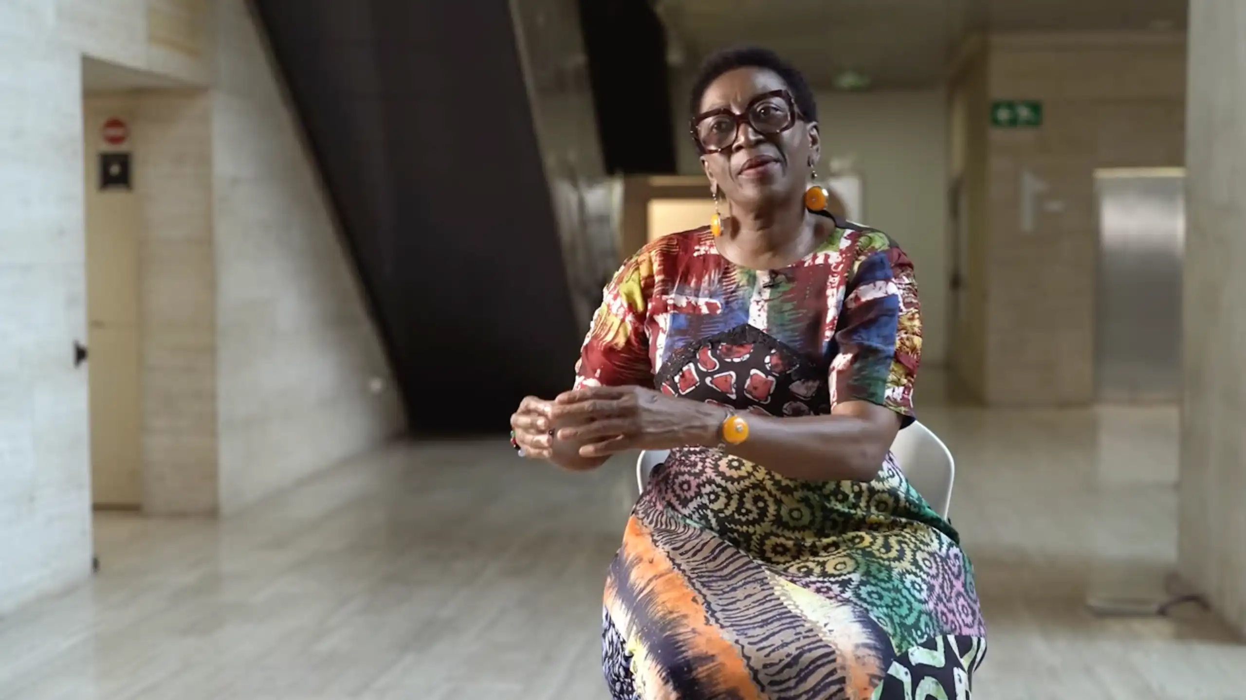 An African woman in heavy-rimmed angular glasses and a bright dress gesures as she sits talking in a chair in a modern architectural space.