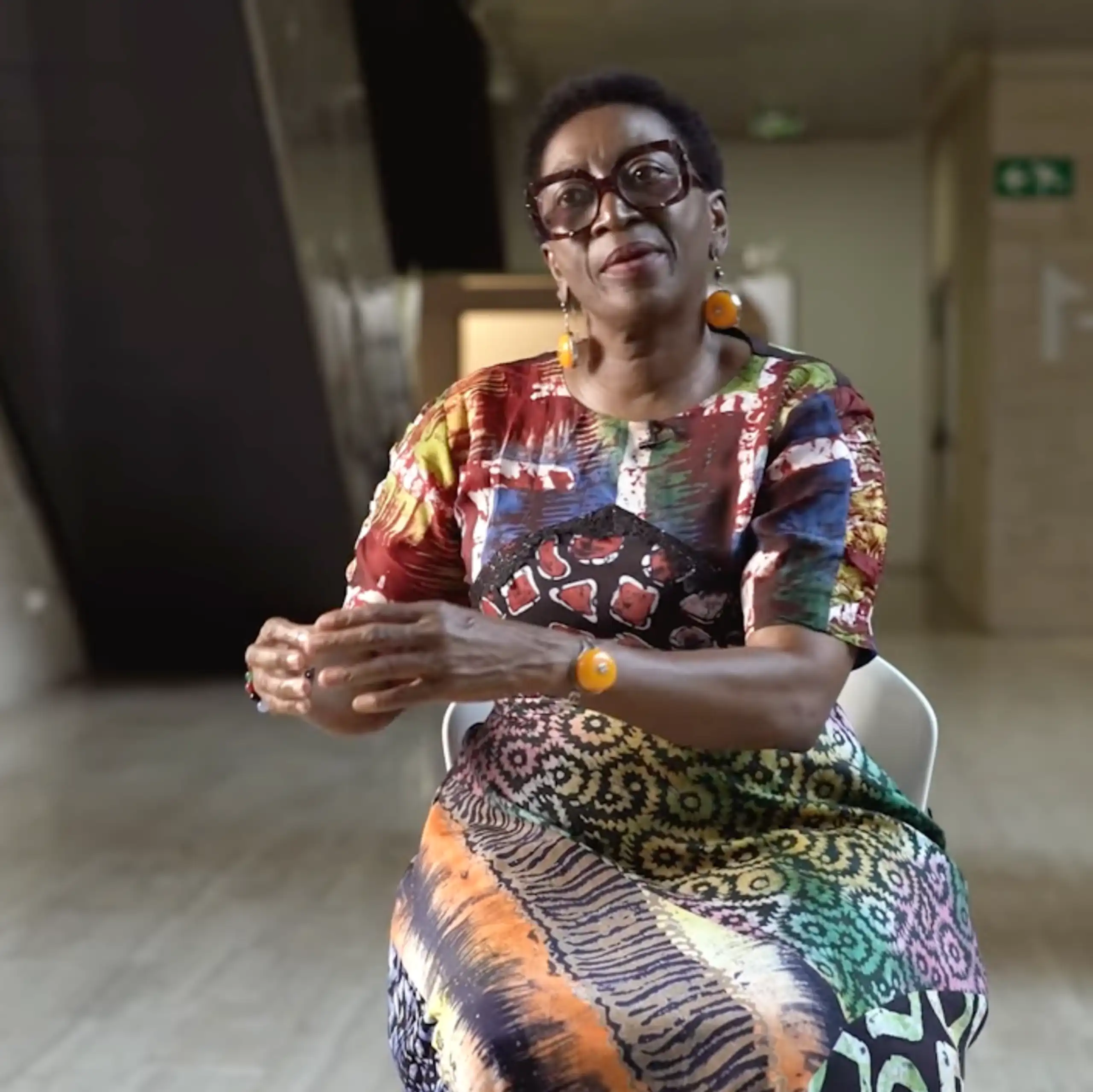 An African woman in heavy-rimmed angular glasses and a bright dress gesures as she sits talking in a chair in a modern architectural space.