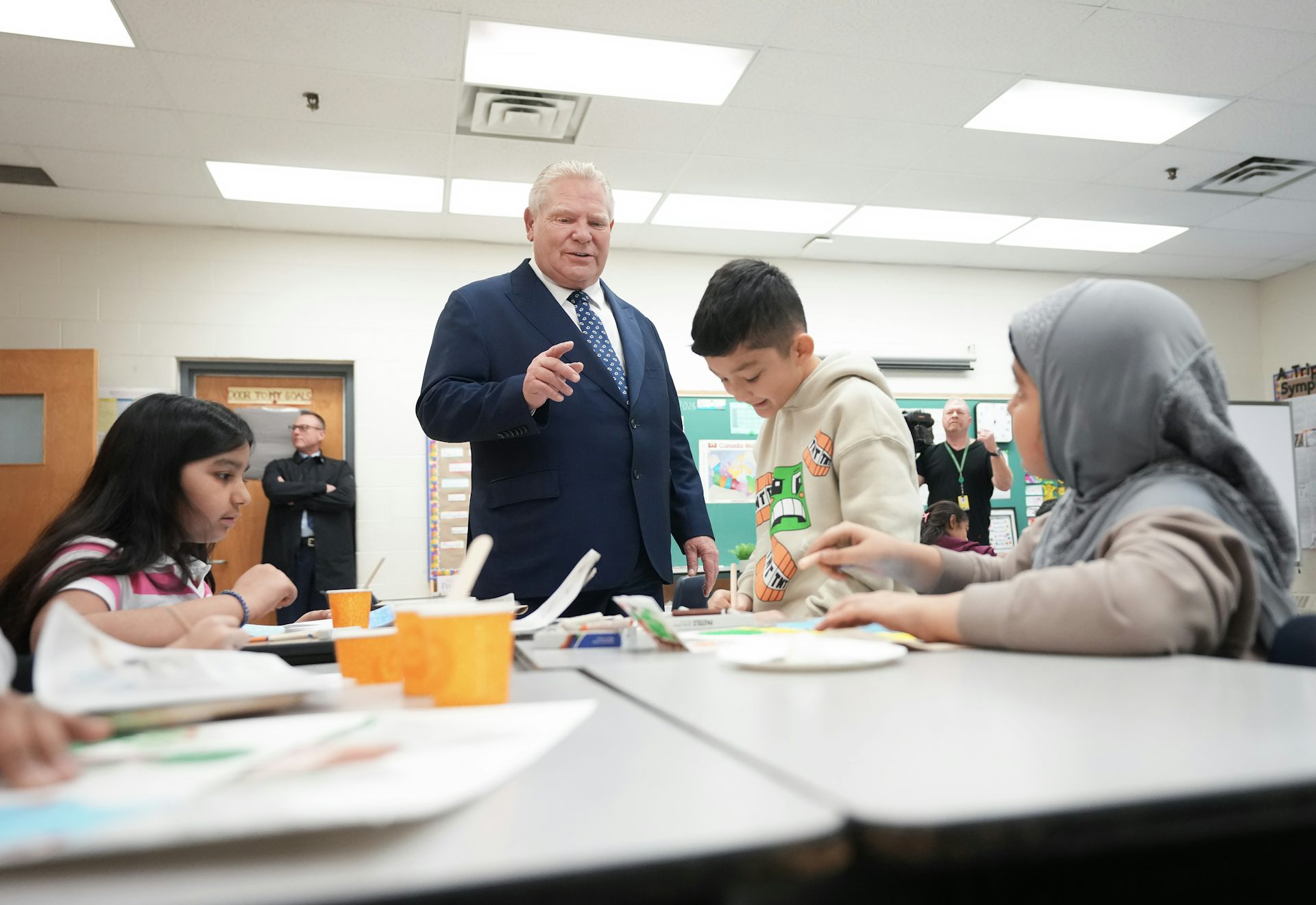 A man in a suit standing in a classroom next to students working at desks.