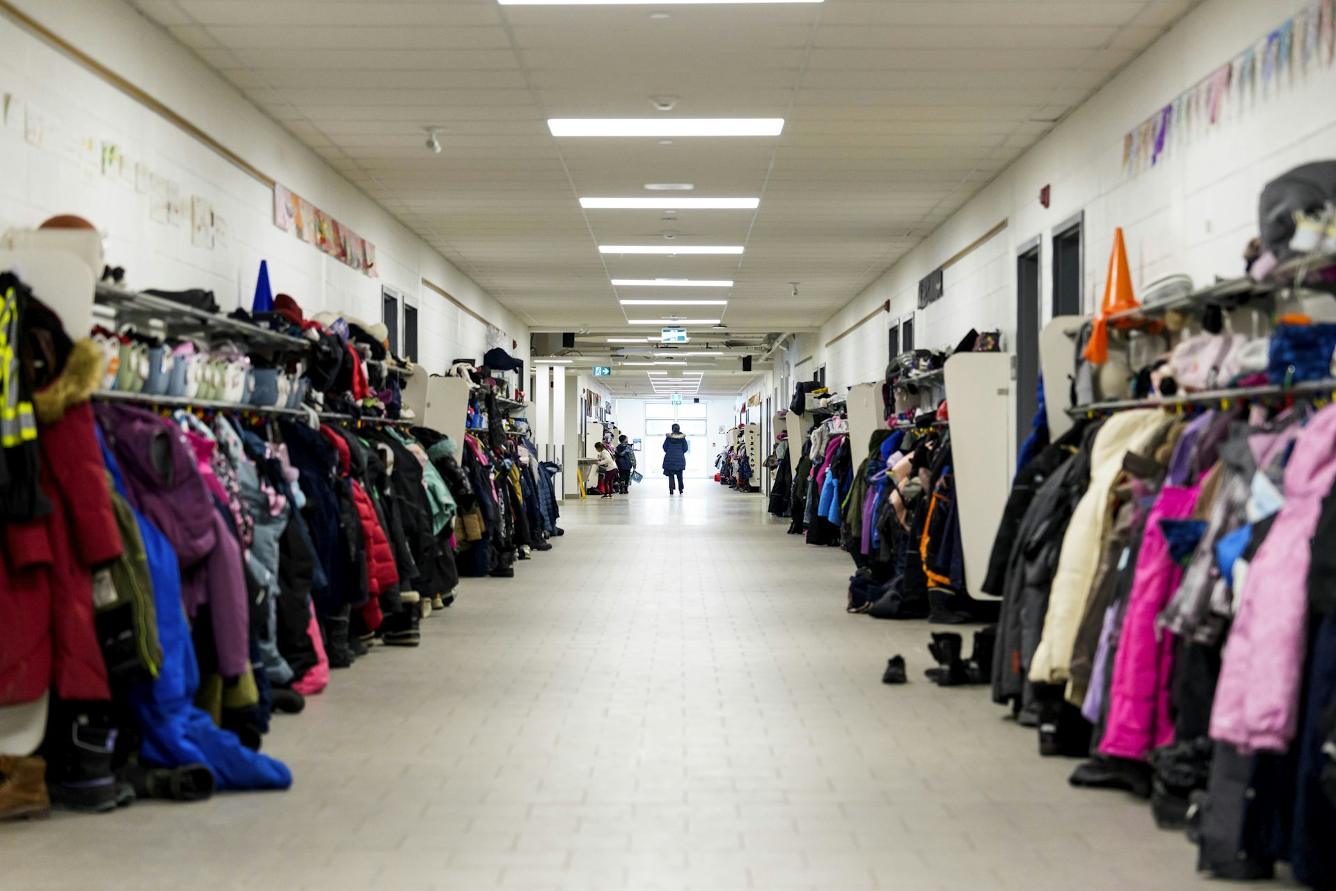A person seen at the far end of a school hallway lined with hanging children's coats and snowpants and an orange pylon.