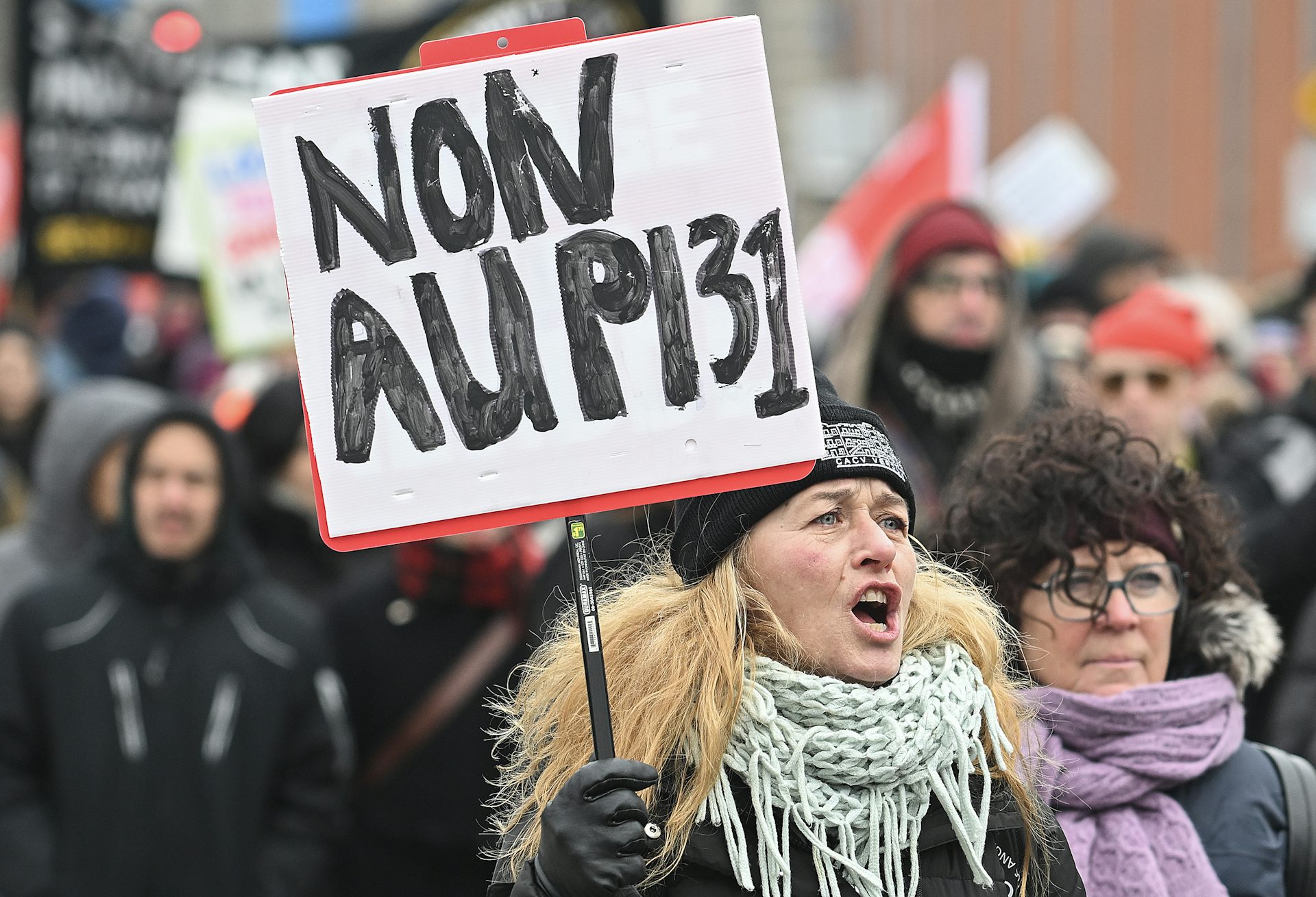 Une femme manifeste parmi une foule en tenant une affiche sur laquelle on peut lire : « Non au PL31 »
