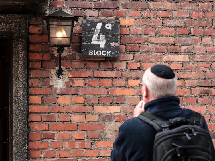 A man with short white hair, a black jacket and backpack and kippah on his head stands in front of a brick wall that says 4 block.