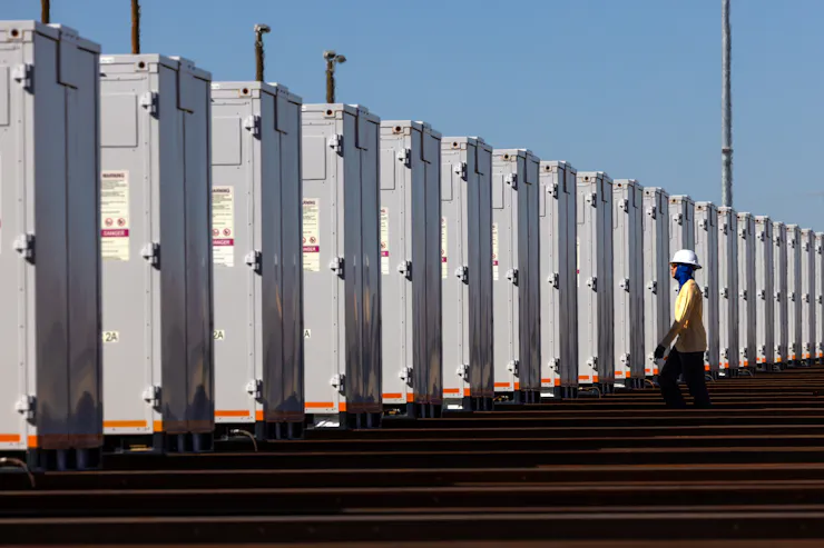 A person wearing reflective clothes and a hard hat stands next to a row of large metal containers.