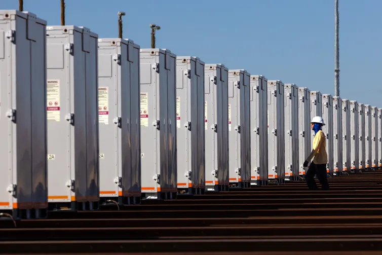 A person wearing reflective clothes and a hard hat stands next to a row of large metal containers.