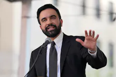 A man in his 30s with dark hair and a beard wearing a suit speaks into a microphone