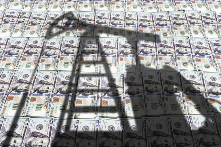 A shadow of an oil pump jack over a spread of hundred-dollar bills.