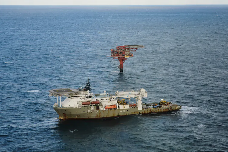 A large platform rises on a pillar out of the ocean, with a ship in the foreground.