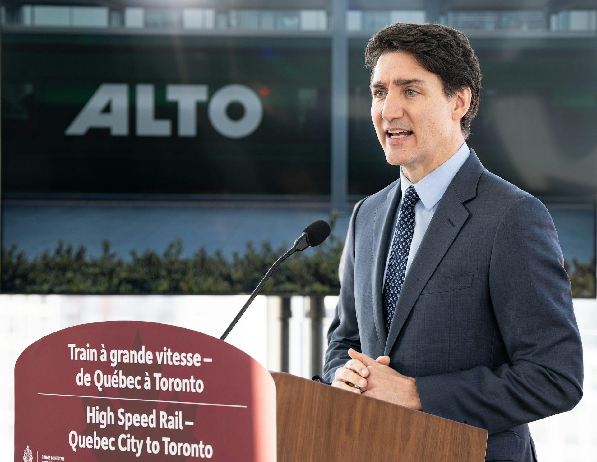 a man in a dark suit speaks at a podium, the word alto behind him