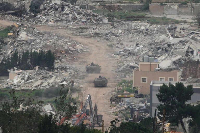 Two tanks drive along a street with destroyed buildings and piles of rubble on either side