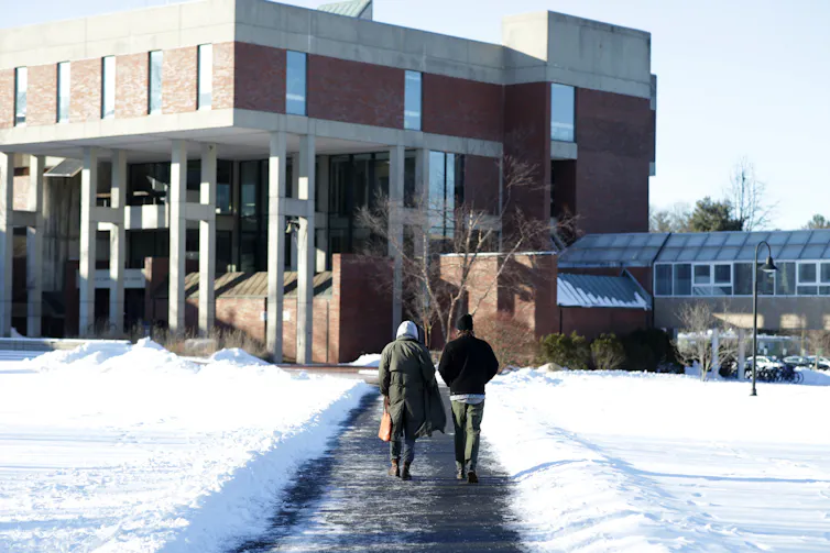 Two people are seen walking down a cleared path towards a large building, with snow all around them.