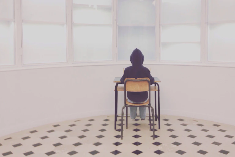 A person wearing a dark hoodie sits at a desk and faces toward a wall in a cartoon image.