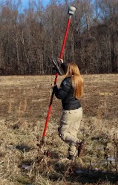 Una mujer sostiene un palo alto mientras camina por un campo con árboles a un lado.