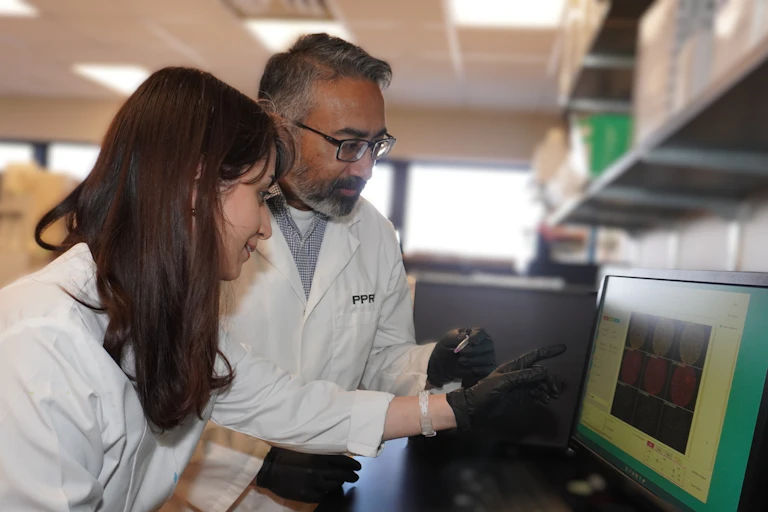 A female and a male researcher in white lab coats examine images on a computer screen.