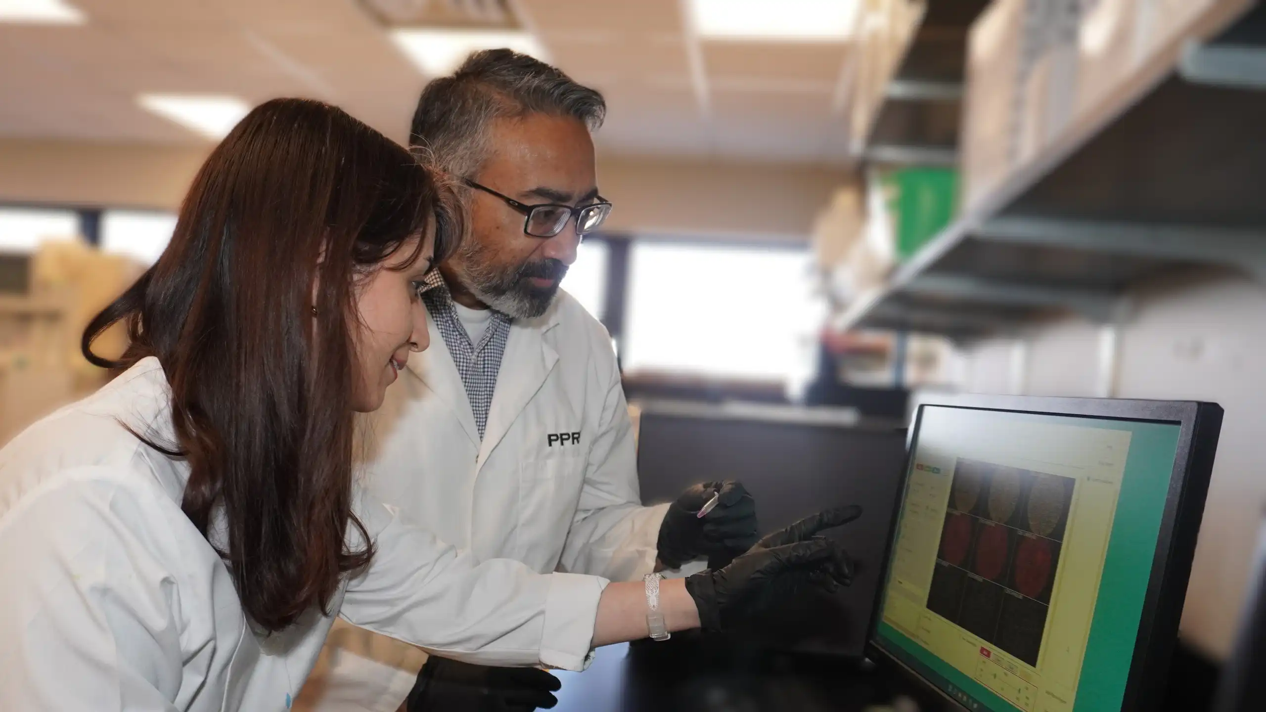 A female and a male researcher in white lab coats examine images on a computer screen.