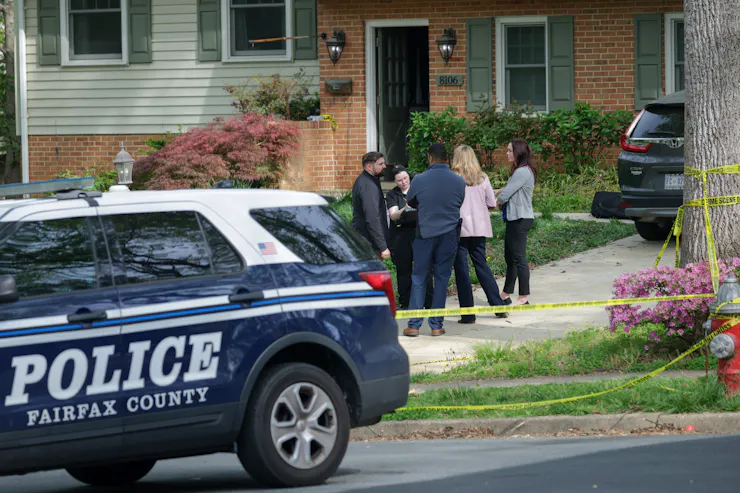 A police car parked on the street and a small group of people standing in front of an open door of a suburban home.