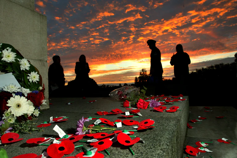 poppies and bouquets strewn on a war memorial with people silhouetted against the dawn light in the background