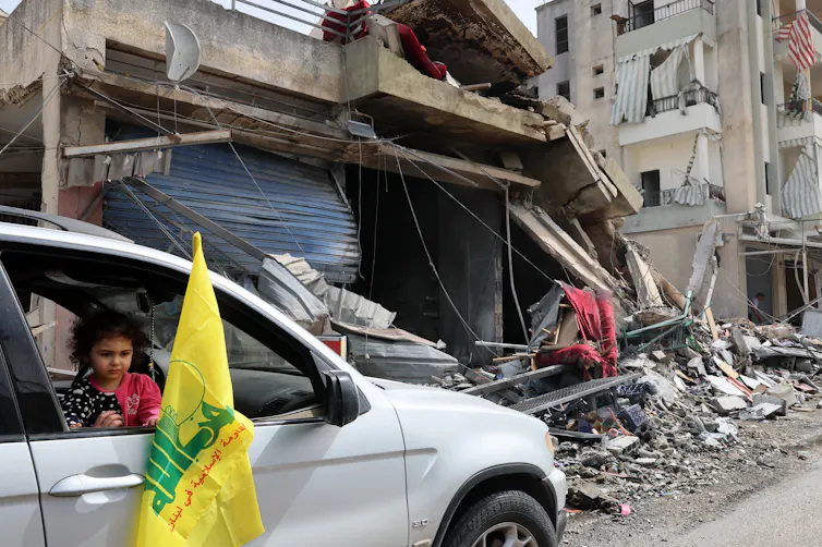 A young girl waves a Hezbollah flag out of the window of her family car as they drive through a destroyed street.