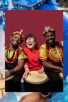 Three people laughing, the woman in the middle plays a drum held on her lap.