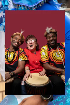 Three people laughing, the woman in the middle plays a drum held on her lap.