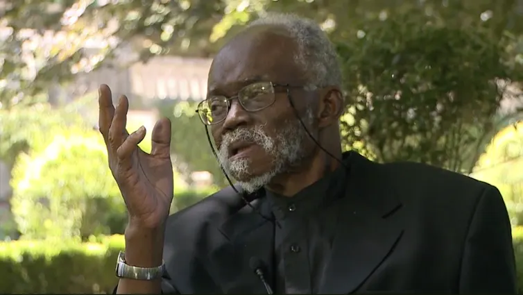 An elder African man with grey hair and spectacles raises a hand as he sits talking in a leafy setting.