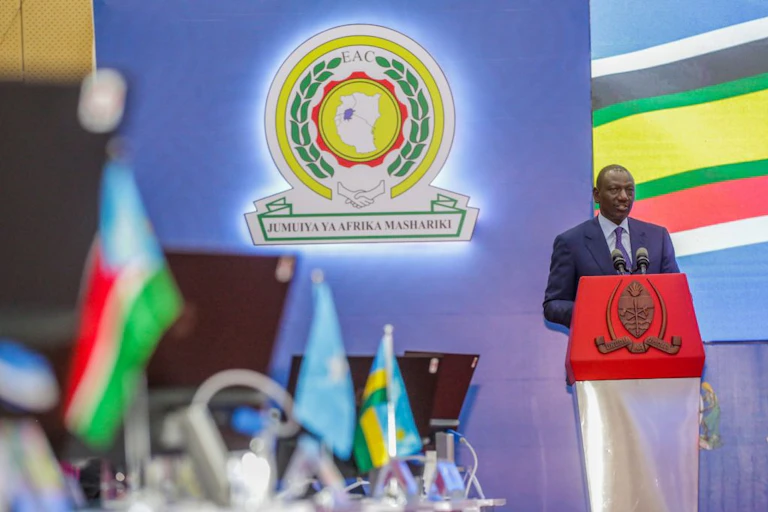 A man speaking at a podium with several flags lined up on a desk in the foreground