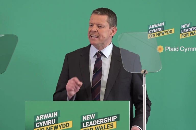 Rhun ap Iorwerth standing behind a green lectern against a green background.