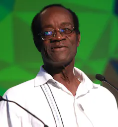 A balding African man in glasses sits in front of a microphone in a casual white shirt.