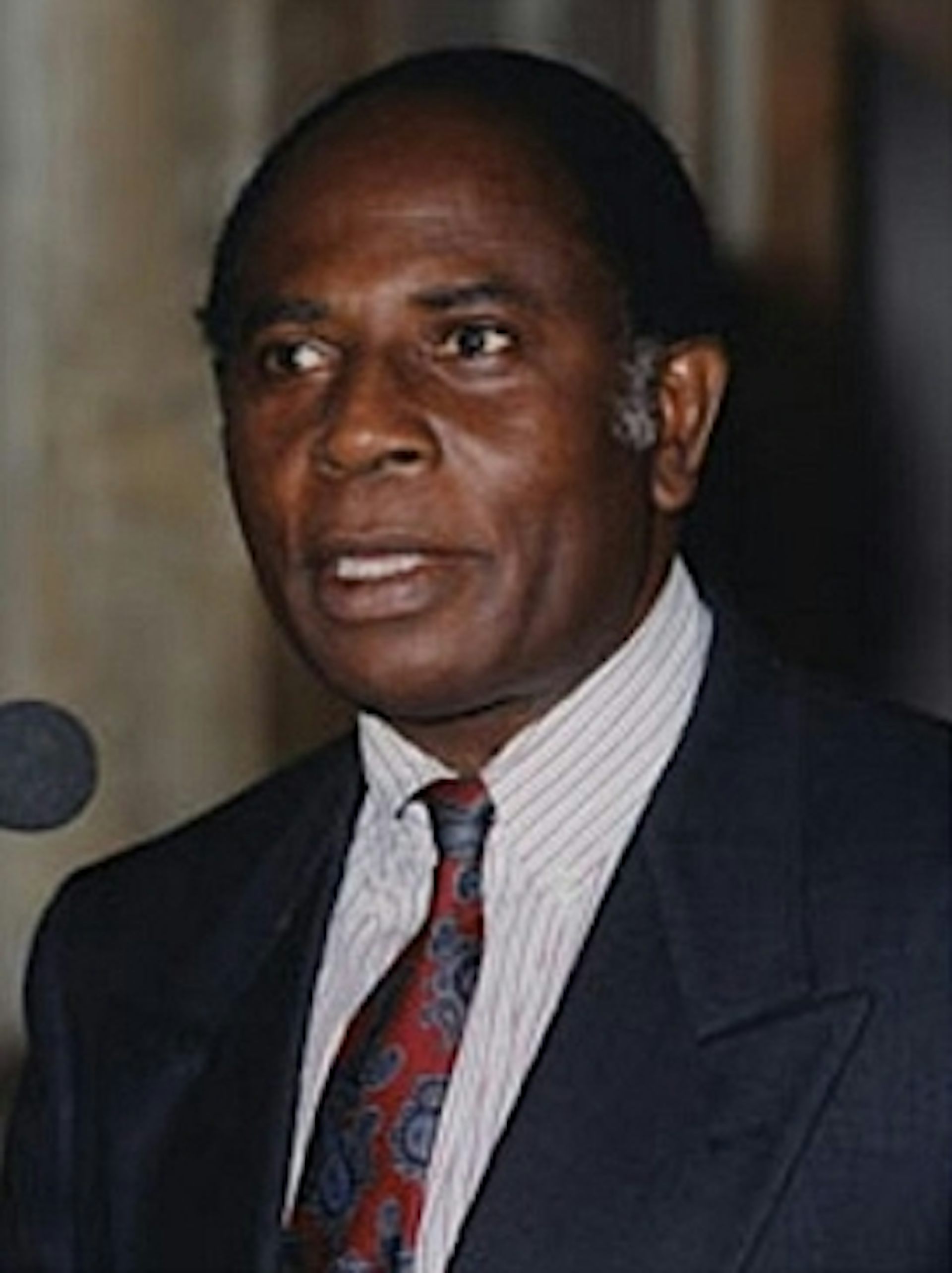An African man in a suit and tie stands at a microphone.