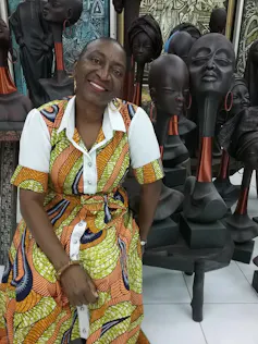 An African woman with short hair sits smiling in a chair in front of African wood carvings.